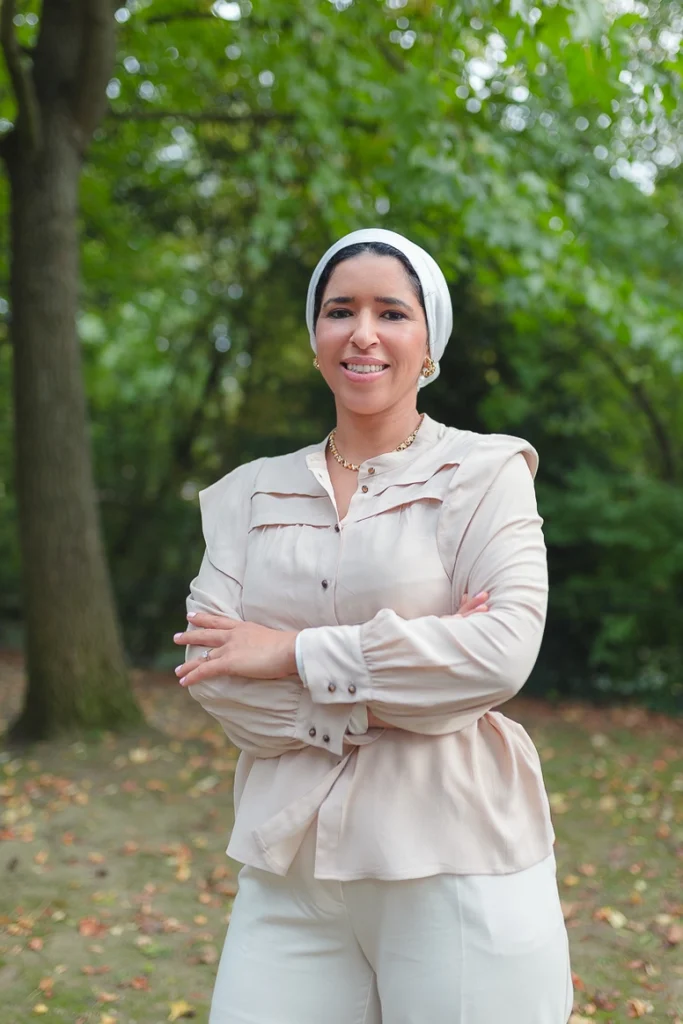 Asma Dana en plan américain, les bras croisé, souriante et l'air décidé. En arrière-plan, une pelouse, le tronc et le feuillage d'un grand arbre feuillu.