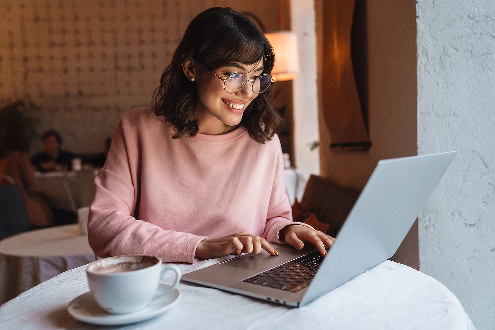 Jeune femme assise seule à une table ronde, en plan poitrine, souriant et tapant sur un ordinateur, avec une tasse de café. 